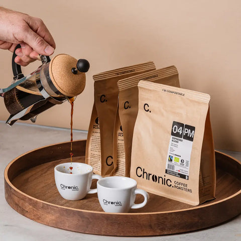 3 paquets de café Chronic sur un plateau avec une french press qui fait couler café dans 2 tasses de café